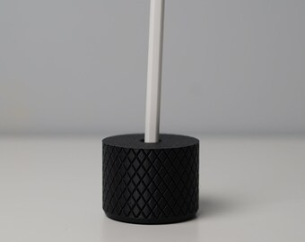 May include: A black, diamond-patterned stand holds a white Apple Pencil.
