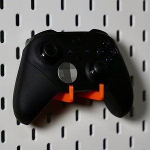 Puede incluir: Un mando Xbox negro con soportes de montaje naranja. El mando está montado en una pared de tablero de clavijas blanco.