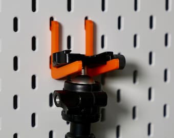 May include: A black tripod head with a ball joint is mounted on a pegboard using two orange plastic hooks.