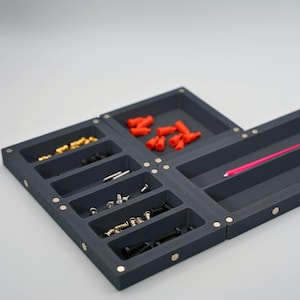 May include: A dark grey, modular organiser with multiple compartments, each holding various small hardware items. The compartments contain items in gold, black, and silver, along with red rubber caps and a pink rod. The organiser is designed for efficient storage.