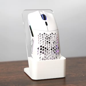 Puede incluir: Un ratón gaming blanco con diseño de panal y luces RGB, exhibido en un soporte de acrílico transparente. El ratón tiene una forma elegante y ergonómica, con rueda de desplazamiento y botones. El soporte es blanco y rectangular.
