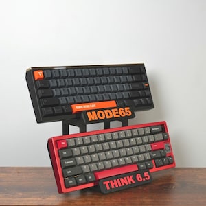 May include: Two mechanical keyboards, one black and one red, are displayed. The black keyboard has orange accents and the text "MODE65". The red keyboard has grey keys and the text "THINK 6.5". They are on a wooden surface.