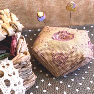 May include: A hexagonal pincushion with a floral pattern in shades of pink and brown. The pincushion is made of fabric and has two decorative pins with colorful tops.