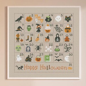 May include: A framed cross-stitch design with a beige background and a white border. The design features a grid of 31 squares, each containing a different Halloween-themed icon, such as a black cat, a ghost, a pumpkin, and a witch's hat. The numbers 1 through 31 are printed in the squares, and the text 'Happy Halloween' is printed below the grid. The frame is hanging on a light beige wall, and a white stool is in the foreground.