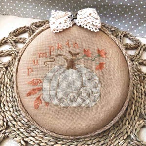 May include: A cross-stitched pumpkin design with the words "pumpkin" and "patch" stitched in a circular frame. The pumpkin is white, with orange leaves and a brown stem. A white lace bow is at the top.