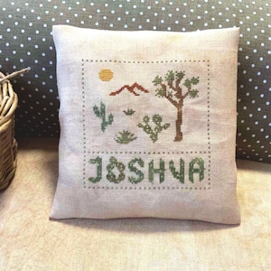 May include: A beige cushion with a cross-stitch design featuring a Joshua Tree, mountains, a sun, and the word "JOSHUA".