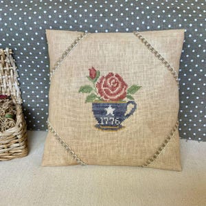 May include: A square, beige pillow with a cross-stitched design. The design features a red rose in a blue teacup with a white star and the year "1776". The pillow is trimmed with a decorative gold cord.