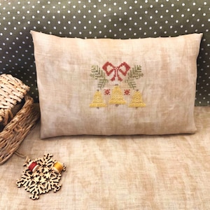 Op de afbeelding: Een beige linnen kussen met een handgestikt ontwerp van drie gele bellen met een rode strik en groene bladeren. Het kussen ligt op een beige stof met witte stippen.