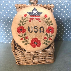Puede incluir: Un diseño de punto de cruz redondo beige con la palabra "USA" en azul marino, rodeado de amapolas rojas y hojas verdes. Encima de la palabra hay un diseño de estrella y rayas. El punto de cruz está en una pequeña cesta tejida.