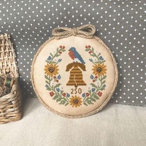 Könnte beinhalten: Ein runder Kreuzstich-Stickrahmen mit einem Glockenmotiv und der Zahl 250. Die Glocke ist von einem floralen Kranz mit Sonnenblumen, blauen Blumen und roten Akzenten umgeben. Ein blau-roter Vogel sitzt auf der Glocke. Der Rahmen ist mit Bindfaden verziert und hat eine Bindfaden-Schleife.
