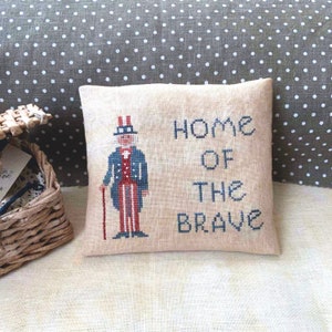 May include: A beige pillow with a cross-stitch design featuring Uncle Sam, a red, white, and blue figure, and the text "Home of the Brave".