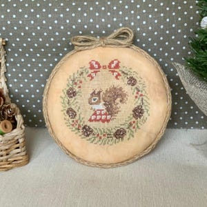 May include: A round, cross-stitched ornament featuring a squirrel wearing a red and white checkered outfit, surrounded by a wreath of pinecones and berries. The ornament has a jute rope border and a jute bow at the top. The background is a light beige fabric.