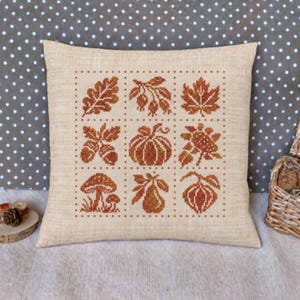 May include: A square beige throw pillow featuring a cross-stitch grid pattern. The design displays nine distinct autumn-themed motifs in shades of orange and brown, including leaves, acorns, a pumpkin, mushrooms, and a pear, all on a linen-like fabric.