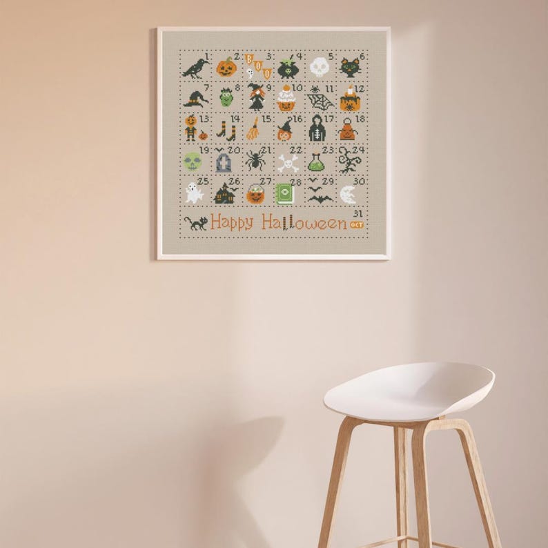 May include: A framed cross-stitch design with a beige background and a white border. The design features a grid of 31 squares, each containing a different Halloween-themed icon, such as a black cat, a ghost, a pumpkin, and a witch's hat. The numbers 1 through 31 are printed in the squares, and the text 'Happy Halloween' is printed below the grid. The frame is hanging on a light beige wall, and a white stool is in the foreground.