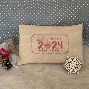 May include: A rectangular beige pillow with red cross-stitch embroidery that reads "HAPPY 2024 NEW YEAR." The pillow is decorated with festive ornaments. A wooden snowflake and a pine cone with a red flower and ribbon are next to the pillow.