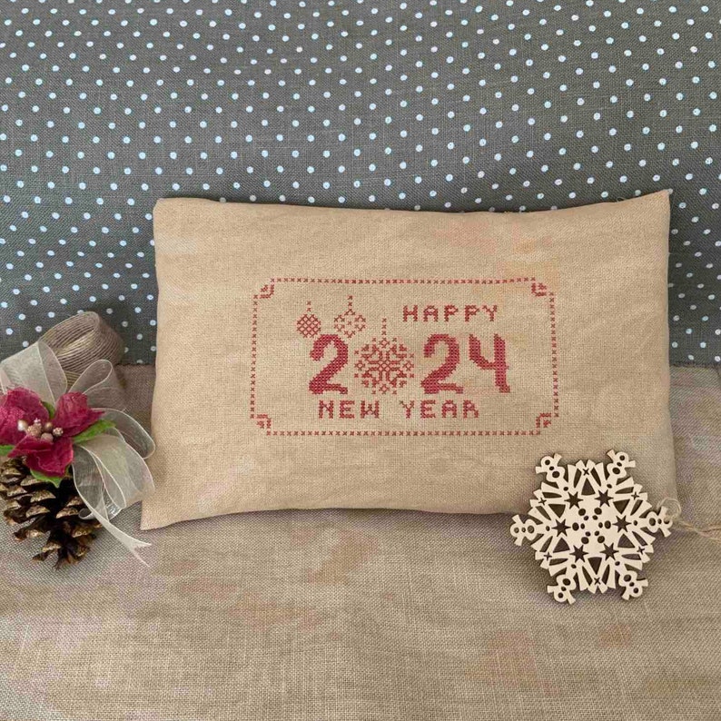 May include: A rectangular beige pillow with red cross-stitch embroidery that reads "HAPPY 2024 NEW YEAR." The pillow is decorated with festive ornaments. A wooden snowflake and a pine cone with a red flower and ribbon are next to the pillow.