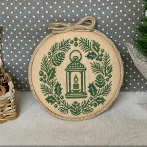 May include: A circular embroidery hoop containing a cross-stitch design on beige fabric. The design features a green lantern surrounded by a wreath of pine branches, pinecones, and holly leaves. A jute twine bow is attached to the top of the hoop.
