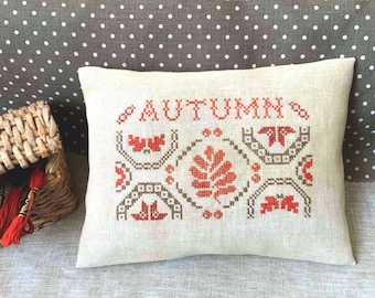 May include: A beige cushion with the word "AUTUMN" embroidered in brown thread. The cushion has a cross-stitch design with brown and orange thread, featuring leaves and other autumnal motifs.