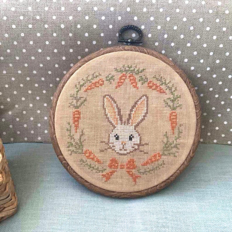 May include: A white bunny face embroidered on a beige fabric with a green wreath of carrots around it. The embroidery is framed in a brown wooden hoop.