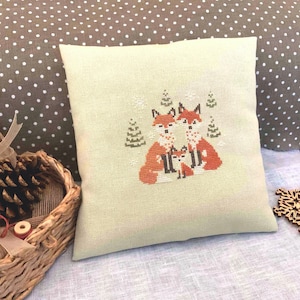 May include: A beige pillow with a cross-stitch design of a fox family in a snowy forest. The foxes are orange and brown with black eyes and noses. The forest is green and white.