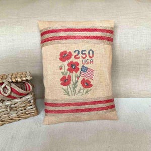 May include: A rectangular beige burlap pillow with red stripes and embroidered details. The pillow features red poppies, the number "250", the word "USA", and a small American flag. A woven basket with red and beige accents is visible on the left.