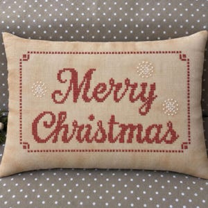 May include: A rectangular beige throw pillow featuring the text Merry Christmas embroidered in red cross-stitch. The design includes a red cross-stitch border and small white cross-stitch snowflake accents on a light tan fabric surface.