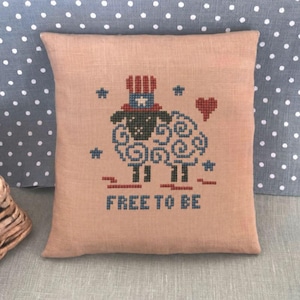 Può includere: Un cuscino quadrato color marrone chiaro con un ricamo a punto croce raffigurante una pecora che indossa un cilindro rosso, bianco e blu. Il disegno include piccole stelle, un cuore rosso e la scritta "FREE TO BE" ricamata in filo blu.