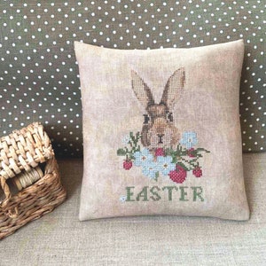 K&ouml;nnte beinhalten: Ein beigefarbenes Kissen mit einer Kreuzstich-Stickerei eines braunen Kaninchens, umgeben von Blumen und dem Wort "Easter".