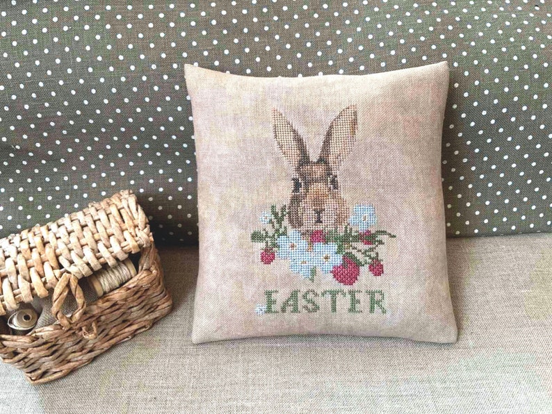 K&ouml;nnte beinhalten: Ein beigefarbenes Kissen mit einer Kreuzstich-Stickerei eines braunen Kaninchens, umgeben von Blumen und dem Wort "Easter".