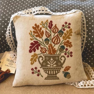 May include: A square beige fabric pillow featuring a cross-stitch design of a decorative urn filled with autumn leaves, acorns, and berries. The pillow is edged with white lace trim and includes a tag labeled Classic Colorworks Fallen Leaves.