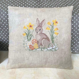 May include: A brown bunny rabbit sitting in a field of yellow daffodils and green grass. The bunny is surrounded by colorful Easter eggs.