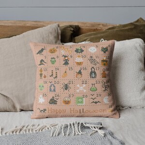 K&ouml;nnte beinhalten: Ein rosa Kissen mit einem Halloween-Kreuzstich-Design. Das Design zeigt 31 Quadrate, jedes mit einem anderen Halloween-Symbol, wie einem K&uuml;rbis, einem Geist, einer Hexe, einer Spinne und einer Fledermaus. Die Zahlen 1 bis 31 sind in den Quadraten gedruckt. Das Kissen tr&auml;gt die Aufschrift "Happy Halloween".
