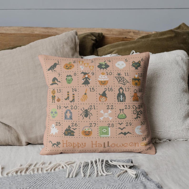 K&ouml;nnte beinhalten: Ein rosa Kissen mit einem Halloween-Kreuzstich-Design. Das Design zeigt 31 Quadrate, jedes mit einem anderen Halloween-Symbol, wie einem K&uuml;rbis, einem Geist, einer Hexe, einer Spinne und einer Fledermaus. Die Zahlen 1 bis 31 sind in den Quadraten gedruckt. Das Kissen tr&auml;gt die Aufschrift "Happy Halloween".