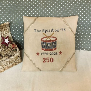 May include: A beige square pillow with the text "The Spirit of '76" and a cross-stitched drum design in red, white, and blue. The pillow also features the dates "1776-2026" and the number "250". A decorative trim borders the pillow.
