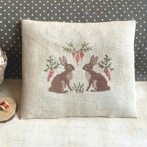 Puede incluir: Una almohada de lino beige con un diseño bordado de dos conejos marrones que se miran. Los conejos están rodeados de follaje verde y zanahorias naranjas.
