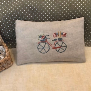 May include: A beige linen pillow with a cross-stitch design of a red bicycle with a basket of flowers and two American flags.