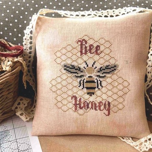 May include: A beige linen cushion with a cross-stitch design of a bee in a honeycomb with the text "Bee Honey". The cushion is trimmed with white lace.
