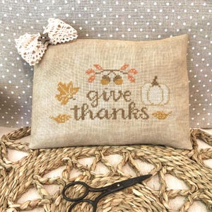 May include: A beige rectangular pillow with the words "give thanks" embroidered in brown, along with fall-themed images. A white lace bow is attached to the pillow. A pair of black scissors rests on a woven mat.