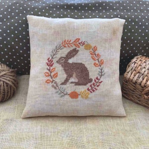 May include: A beige linen pillow with an embroidered design of a brown rabbit sitting inside a wreath of autumn leaves. The wreath is made up of orange, yellow, and red leaves.