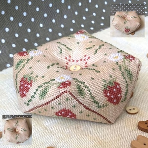 May include: A beige fabric pincushion with a square shape and a button in the center. The pincushion is decorated with embroidered strawberries, leaves, and flowers.