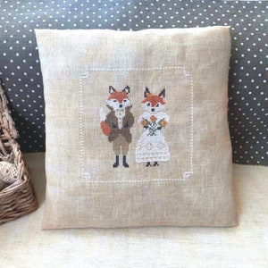 May include: A beige linen pillow with a cross-stitch design of two foxes. The fox on the left is wearing a brown jacket and black pants. The fox on the right is wearing a white dress and holding a bouquet of flowers.
