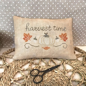 Può includere: Un cuscino rettangolare beige con le parole "harvest time" ricamate. Il design presenta una zucca, foglie e ghiande a punto croce arancioni e verdi. Un paio di forbici nere poggia su un tappetino intrecciato, con uno sfondo a pois.
