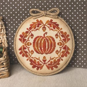 May include: A circular cross-stitch embroidery piece featuring a central orange pumpkin surrounded by a wreath of autumn-colored oak leaves and acorns. The fabric is mounted in a round wooden hoop, finished with a decorative jute rope border and bow.