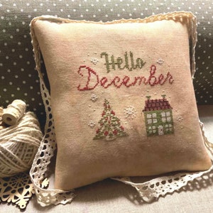 Peut inclure: Un coussin beige avec une bordure en dentelle blanche présente un motif de point de croix d'une maison, d'un sapin de Noël et du texte "Hello December".