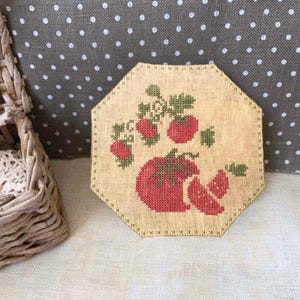May include: A beige, octagonal cross-stitch coaster featuring a red tomato and sliced tomato with green leaves. The design is stitched on a tan fabric with a dotted background and a woven basket in the corner.
