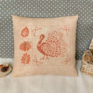 Puede incluir: Cojín cuadrado beige con un diseño de punto de cruz que presenta un pavo real, hojas y una calabaza en tonos naranja. El cojín tiene un fondo neutro y un borde decorativo.