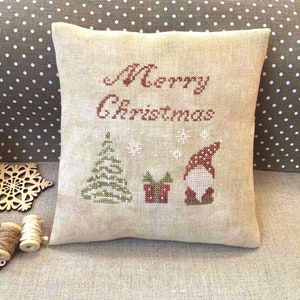 May include: A beige Christmas cushion with cross-stitch embroidery. The words "Merry Christmas" are stitched in red, alongside a Christmas tree, a gift, snowflakes, and a gnome.