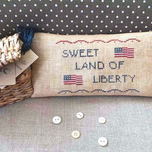 May include: A beige pillow with the words "Sweet Land of Liberty" embroidered in black and red thread. Two small American flags are embroidered on the pillow. There are five white buttons on a beige surface below the pillow.