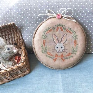 May include: A brown and beige embroidered hoop with a white bunny face surrounded by green leaves and orange carrots. The hoop is tied with a pink ribbon and a pink button.