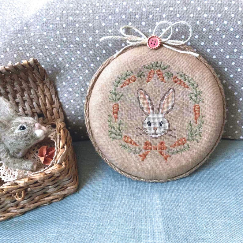 May include: A brown and beige embroidered hoop with a white bunny face surrounded by green leaves and orange carrots. The hoop is tied with a pink ribbon and a pink button.